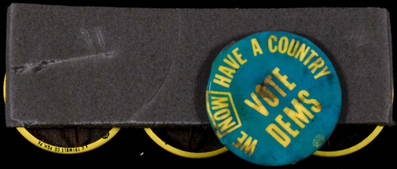 A blue badge mounted on grey foam with the slogan in yellow text 'We now have a country - vote Dems'.