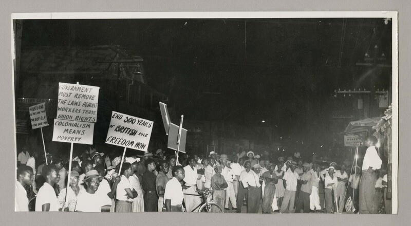 The photograph captures a crowd of people holding up protest signs with a speaker addressing them to the right. The slogans include 'End 300 years of British rule - freedom now!', 'Government must remove the laws against workers trade union rights' and 'Colonialism means poverty'.