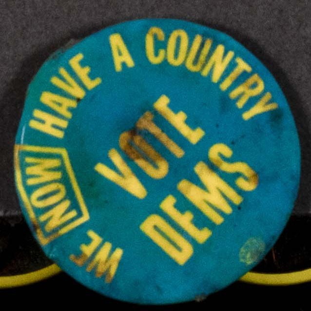 A blue badge with the slogan in yellow text 'We now have a country - vote Dems'.