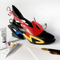 An artistic high-heeled shoe with bold colours and abstract shapes, displayed next to paint tubes and a brush.