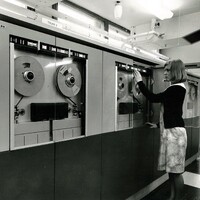 Black-and-white photo of a woman operating a large reel-to-reel computer tape machine in a data centre.