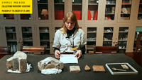 Immie Meade sits at table and reads from script. On the table in front of her are six archaeological objects.