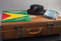 A brown suitcase lying flat. On top of it (left to right) there are a vinyl with the flag of Guyana, a pork pie hat and a stack of airmail letters.