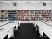 A modern library with shelves full of books in various colours. There are white tables and chairs in the foreground.